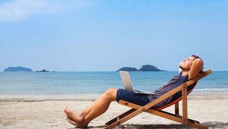 A young man relaxing in a deck chair on the beach with a laptop